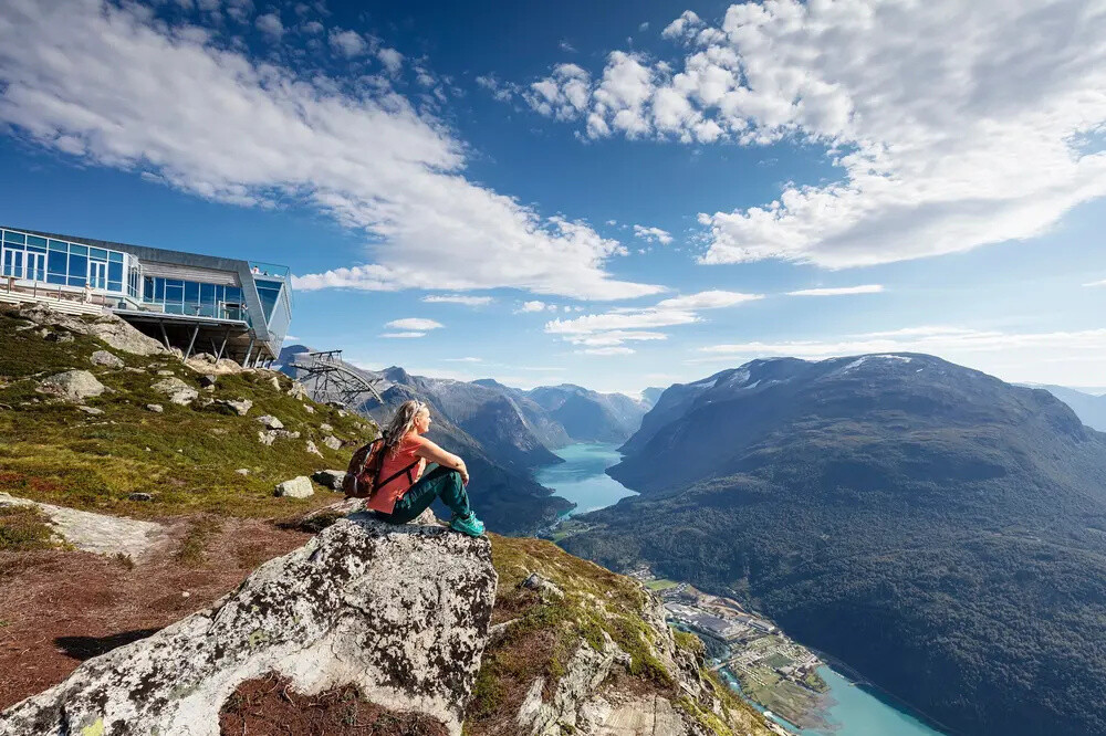 Person sitting on rock, overlooking mountains and fjord, with blue skies.