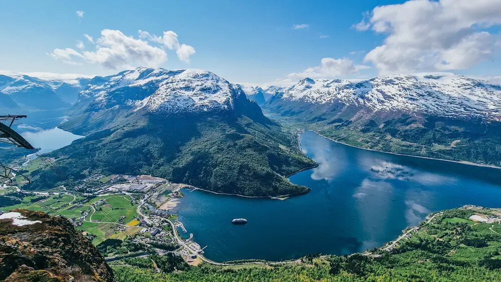 Aerial view of a vast fjord with snow-capped mountains and lush greenery under a clear blue sky.