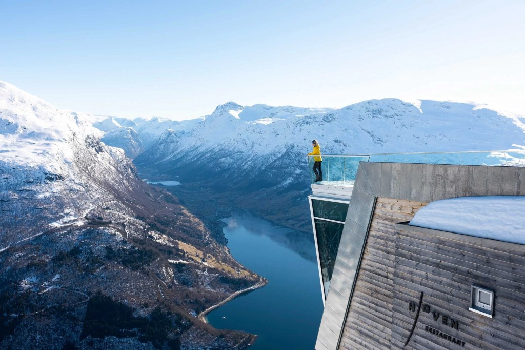 Person standing on glass viewing platform over snowy mountain landscape and fjord.