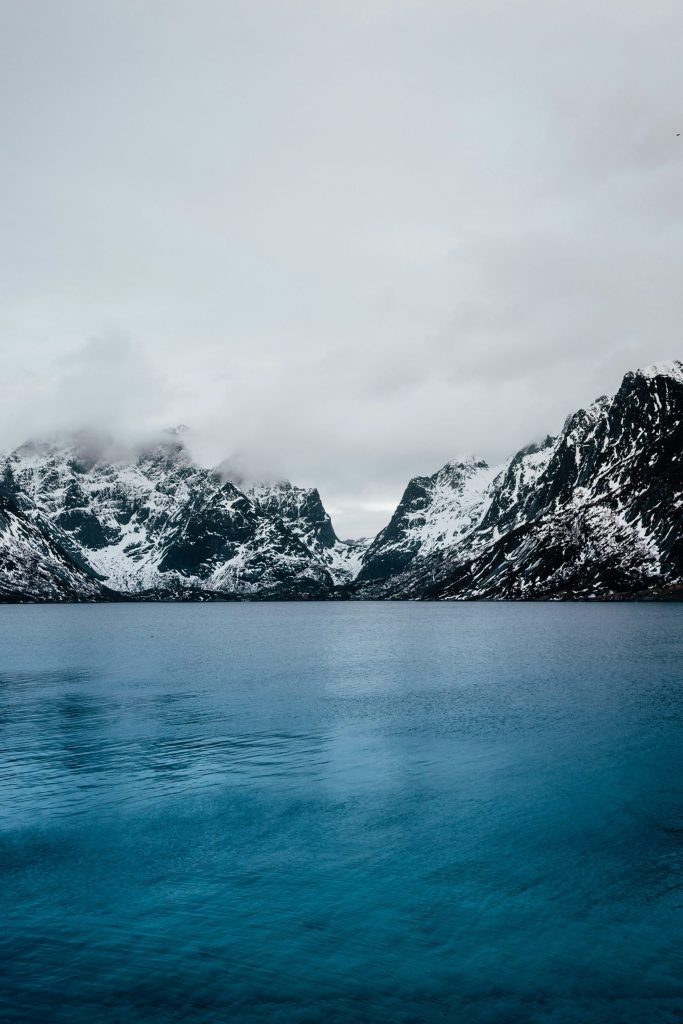Snowy mountains and calm blue lake under cloudy sky.