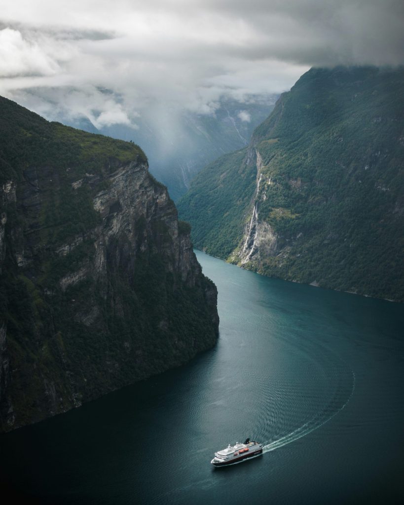 Cruise ship sails through narrow fjord surrounded by steep cliffs under cloudy skies.
