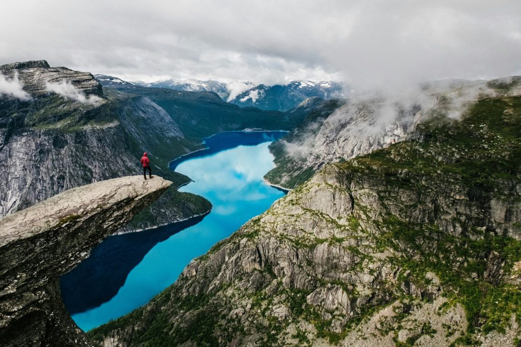 Person standing on a cliff overlooking a blue lake and mountains.