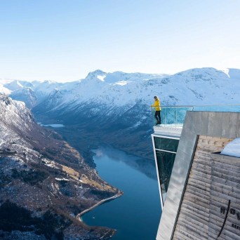 Person standing on glass viewpoint overlooking snowy mountains and fjord.